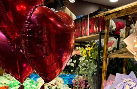 Dükkân içinde kırmızı kalp balonlar, arkada kovalarda mevsim çiçeği ve raflarda hazır buketler