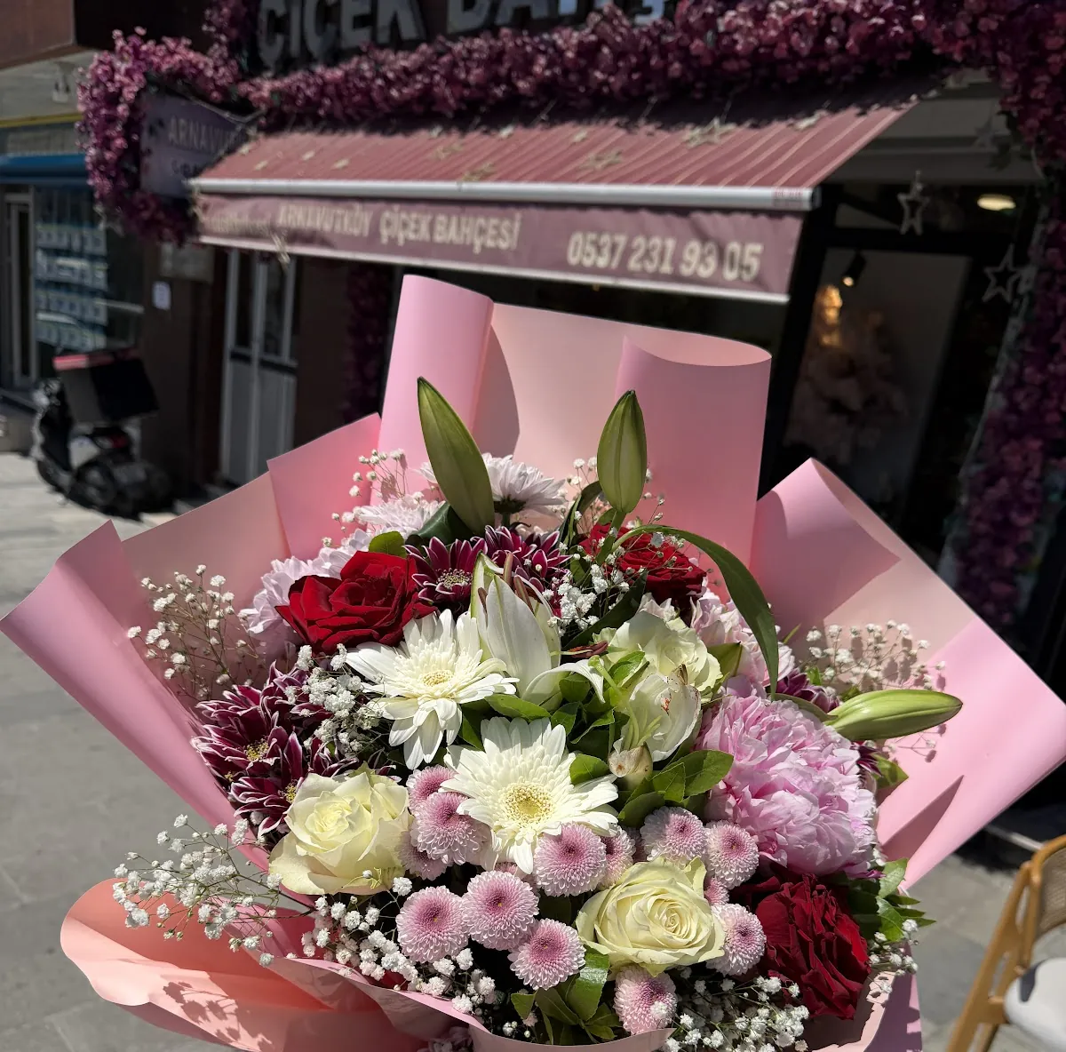 Dükkânın pembe tenteli cephesi önünde tutulan pembe, beyaz ve bordo karışık buket